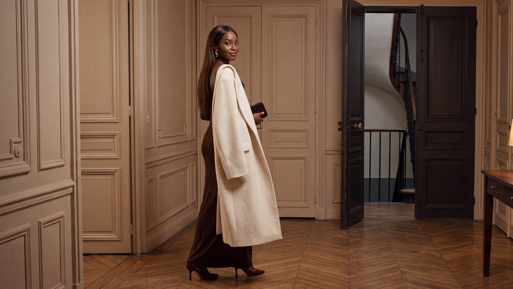Une femme en robe ajustée chocolat, manteau crème sur l'épaule, se retourne en souriant avant de sortir